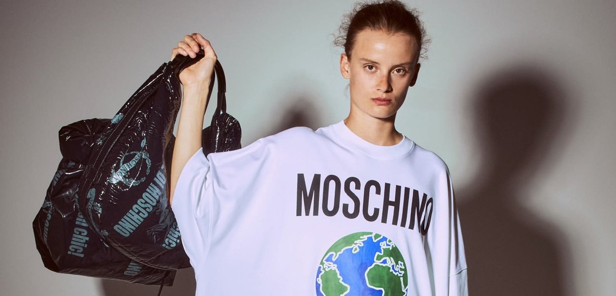 T-shirt bianca oversize con "MOSCHINO" stampato in bold lettere nere e una grafica del globo. Il modello tiene una borsa nera testurizzata con loghi.