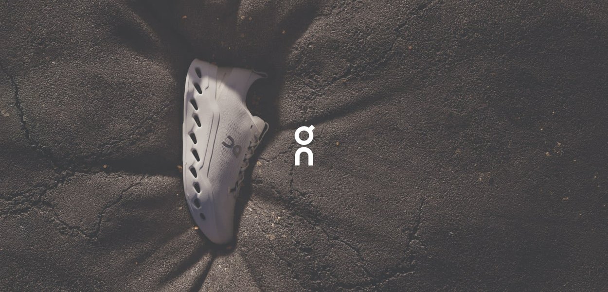 Weiße Sneaker mit einer glatten, abgerundeten Form und seitlichen Belüftungsöffnungen, auf einer strukturierten dunklen Oberfläche präsentiert. Mit Markenlogo an der Seite.