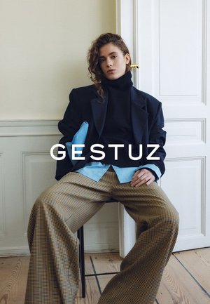 Giovane con capelli ricci seduto su una sedia indossando una giacca nera, camicia blu, dolcevita nero e pantaloni larghi a quadri in una stanza minimalista.