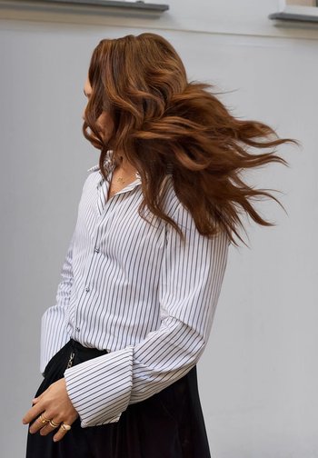 White button-up shirt with black vertical stripes, oversized cuffs, black skirt, long wavy brown hair, against a light gray background.