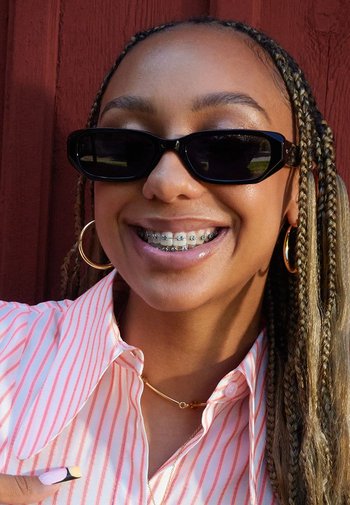 Zwarte zonnebril, roze gestreept shirt, gouden ringoorbellen en beugel. Haar in vlechten, met een lichte glans op de huid. Eenvoudige ketting als accent.