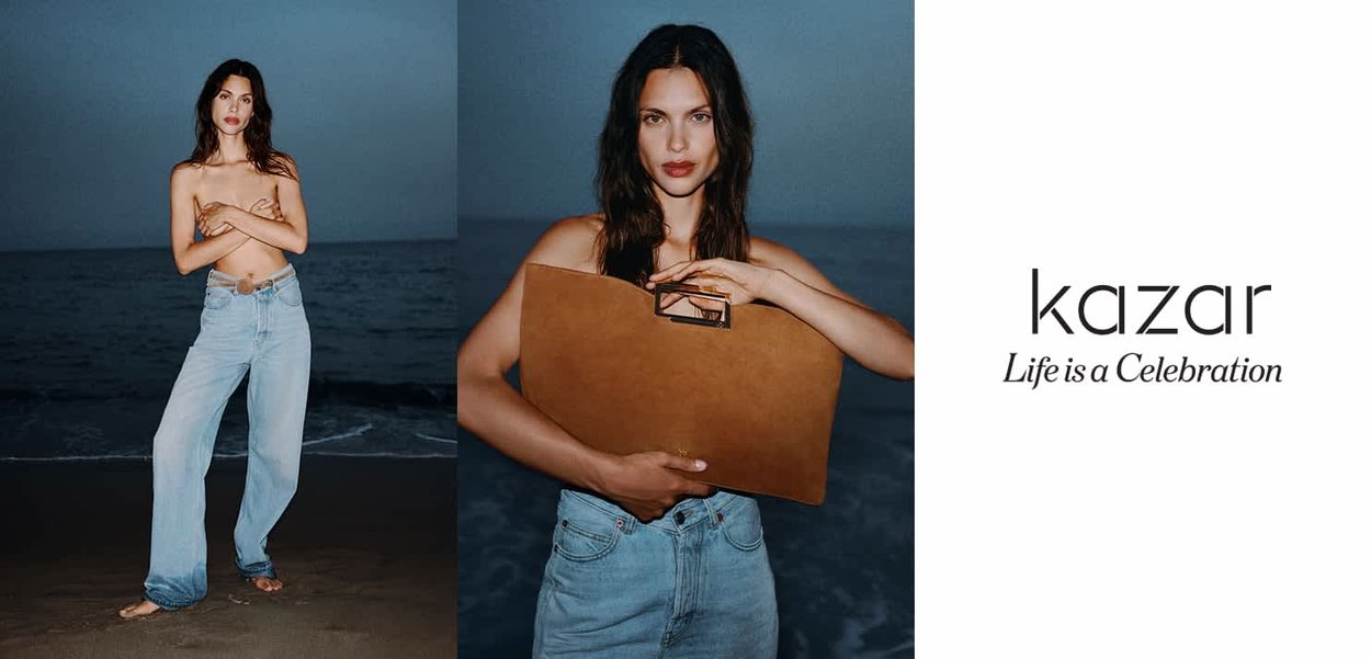 Model holds a tan leather handbag with a gold handle, wearing light blue wide-leg jeans. Background features dark ocean waves and dusk sky.