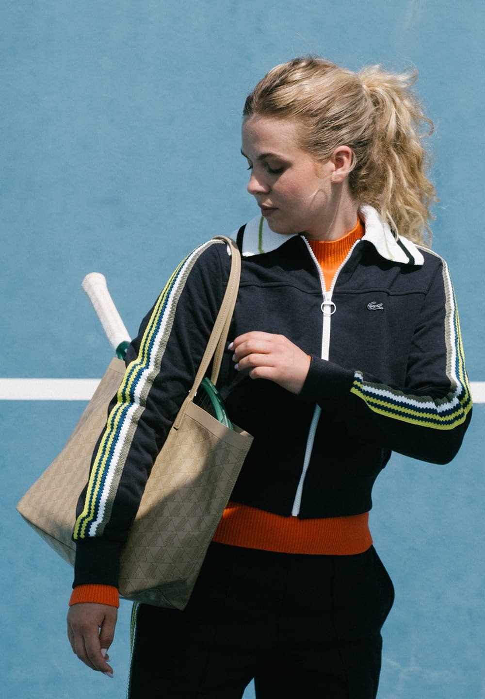 Schwarze Zip-Jacke mit grünen, gelben und weißen Streifen an den Ärmeln, orangefarbener Pullover darunter, beiger Tragetasche und Tennis-Equipment sichtbar.