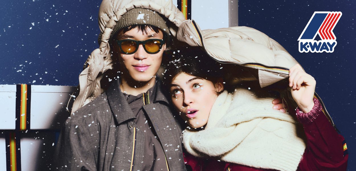 Modella con cappello e guanti neri, giacca a piumino bianca e sciarpa marrone. Fiocchi di neve cadono su uno sfondo blu, sottolineando il tema invernale.