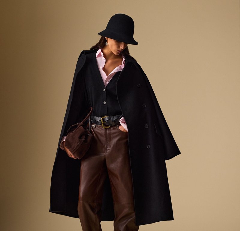 Manteau en laine noir avec de grands boutons, superposé à un gilet noir et une chemise rose pâle. Un pantalon en cuir marron et un sac à main marron foncé complètent le look.