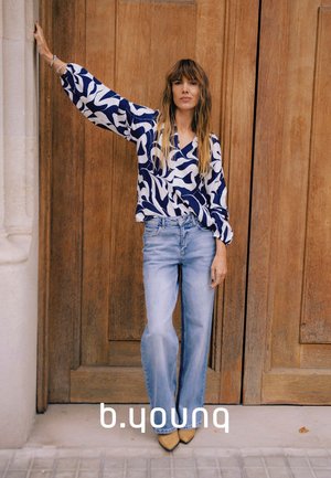 Fließende Bluse mit blau-weißem Muster und langen Ärmeln, kombiniert mit hellblauen Jeans und beigen Stiefeletten, vor Holztüren.