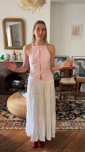 Top halter rosa claro con cortes en forma de bucle y lazos, combinado con una falda larga blanca de volantes y zapatos rojos brillantes. Suelo de madera y decoración al fondo.