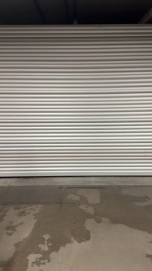 Porte de garage en métal roulant avec des rainures horizontales, finition blanche. Texture lisse, installée au-dessus d'un sol en béton avec des taches d'eau.