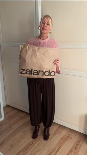 Brown paper bag featuring the word "zalando" in black lettering, being held by an individual dressed in a light pink top and dark, loose-fitting trousers, set against a light-coloured interior.