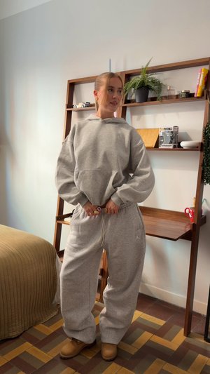 Sudadera y pantalones deportivos grises combinados con botas marrones. El conjunto presenta un ajuste holgado, mangas oversized y un bolsillo canguro en la sudadera con capucha.