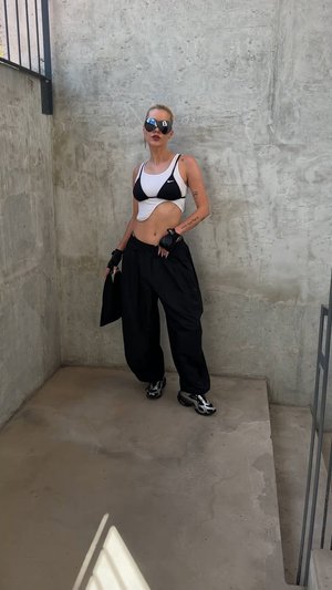 Fekete-fehér sportcrop top, laza fekete nadrág és elegáns fekete sportcipő. Kiegészítők közé tartozik napszemüveg és ujjak nélküli kesztyű. Beton háttér.