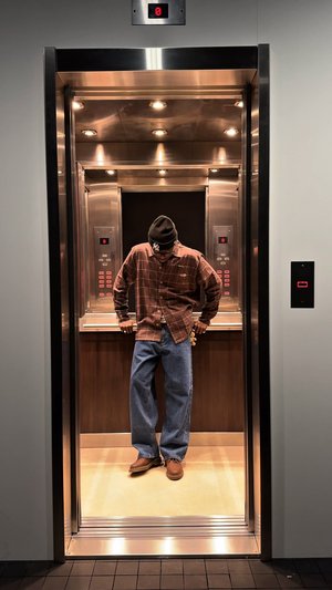 Ascenseur avec parois en acier inoxydable et panneaux en bois. Une personne se tient debout, portant une chemise à carreaux marron, un jean bleu et des chaussures marron.