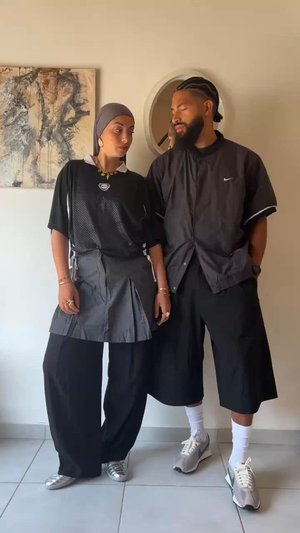 Divi cilvēki stāv tuvu viens otram, ģērbušies melnā sieta sporta krekla, pelēkās svārku un brīviem melniem šortiem. Ievērojami aksesuāri ir rotaslietas un dažādi apavi.