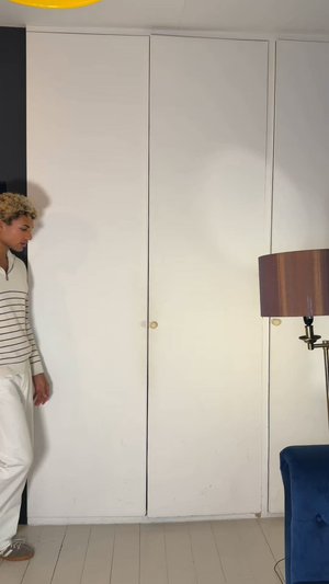 Jeune personne marchant devant des portes d'armoire blanches fermées, avec une lampe sur pied et un fauteuil bleu visibles dans la pièce.