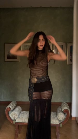 Hosszú sötét hajú nő áll bent, fekete halháló ruhát visel, széles díszes övvel és hosszú francia szoknyával.