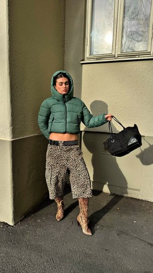 Veste matelassée verte, pantalons larges imprimé léopard, bottines imprimé léopard, sac à main noir avec logo, ventre découvert, debout contre un mur.
