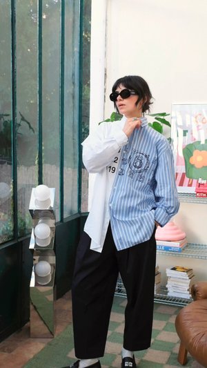 Mėlyna ir balta juostuota marškinėliai su mygtukų uždarymu ir spausdinta dizainu, derinami su laisvais juodais kelnėmis ir juodomis šlepetėmis. Tekstūruotas fonas.