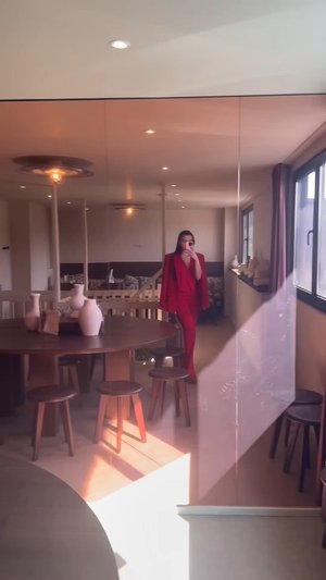 Rode pak met een op maat gemaakte blazer en wijde pijpen, staande naast een ronde houten tafel en verschillende houten krukken in een heldere, moderne interieur.