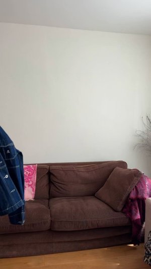 Bruine stoffen bank met twee kussens, textuurstofbekleding en een roze motiefdeken die over de zijkant hangt. Houten vloer op de achtergrond.