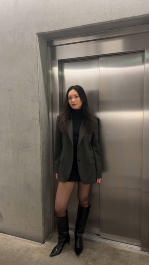 Blazer verde oscuro sobre un cuello de tortuga negro y una minifalda. Botas altas negras. De pie junto a un ascensor plateado, contra una pared de hormigón.