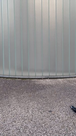 Mur en métal ondulé gris avec des lignes verticales, texture lisse et base arrondie ; surface sombre et texturée en dessous avec de petits cailloux visibles.