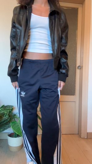 Veste bomber en cuir noir portée sur un débardeur blanc court, associée à un pantalon de survêtement bleu marine avec des bandes blanches sur les côtés et le logo Adidas.