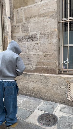Une personne en sweat à capuche gris et en pantalon en denim surdimensionné se tient devant un mur en pierre texturé, à côté d'une fenêtre barrée et d'un couvercle de regard rond.