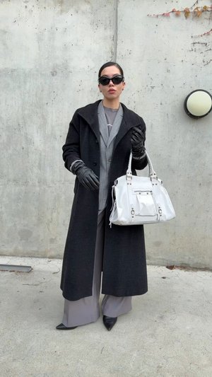 Abrigo negro de lana, blazer gris de gran tamaño, guantes de cuero negro, pantalones grises de pierna ancha, zapatos negros puntiagudos y un bolso blanco con detalles.