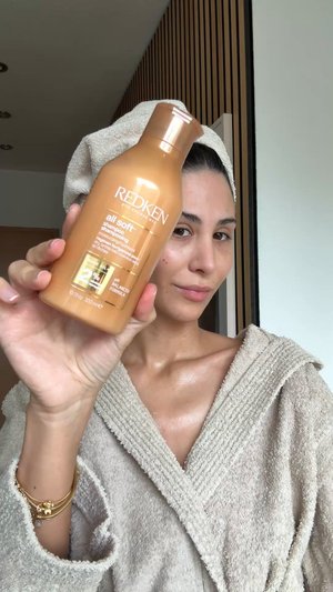 Redken All Soft shampoo in een lichtbruine plastic fles met een pomp, met witte tekst en een gouden accent. Model draagt een badjas van badstof.
