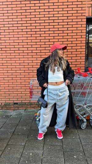 Graue Jogginghose, weißes kurzes Oberteil, schwarzer Mantel und rote Mütze. Accessoires sind eine kleine schwarze Tasche und rosa Turnschuhe. Einkaufswagen im Hintergrund.