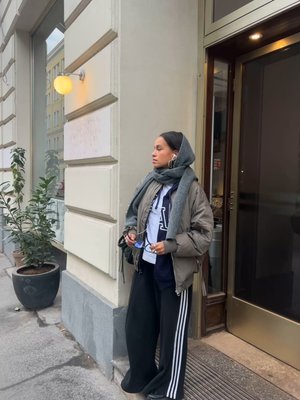 Doudoune grise avec capuche texturée, sweat-shirt bleu foncé, large pantalon noir avec des rayures blanches, et un petit sac noir ; debout près d'une fenêtre.