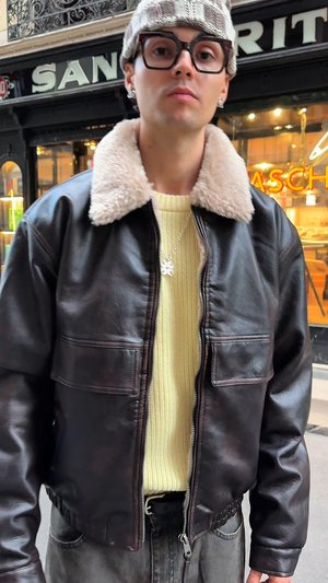 Jeune personne portant de grandes lunettes, un bonnet en tricot gris, une veste en cuir marron foncé avec un col en fourrure, un pull jaune en tricot et un collier argenté dans la rue.