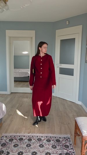 Rotes Strickkleid mit Knopfleiste vorne, langen Ärmeln und plissiertem Rock. Kombiniert mit schwarzen Stiefeln, präsentiert in einem hellen Raum.