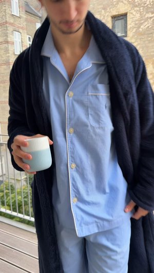 Ensemble de pyjama rayé bleu clair pour homme avec des accents blancs, porté sous un peignoir en polaire bleu marine foncé ; tenant une tasse en céramique avec une finition dégradée.
