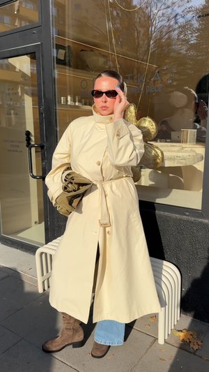 Trench-coat long beige avec ceinture à nouer, fermeture à boutons et manches larges, porté sur un jean bleu et des bottes marron décontractées. Des lunettes de soleil noires complètent le look.