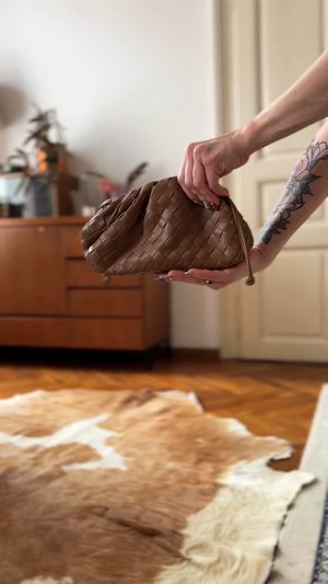 Bolso clutch de cuero marrón tejido con forma redondeada y cierre de cordón, sostenido frente a un fondo de alfombra texturizada.