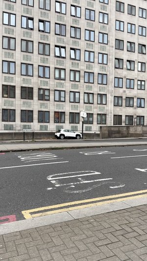 White SUV parked beside a grey concrete building featuring a pattern of dark-framed windows and light panels. The pavement displays a textured design.