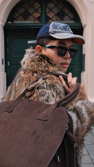 Sac à main en daim marron avec double poignée, présentant un manteau en fourrure aux nuances de brun et crème, porté avec une casquette en jean et des lunettes de soleil.
