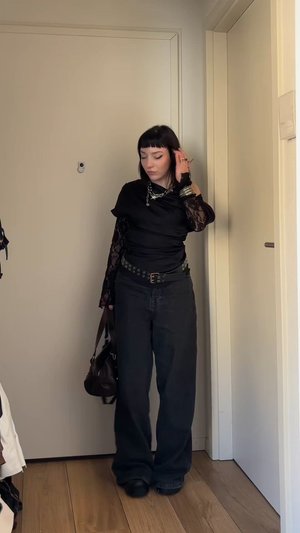 Schwarzes Strickoberteil mit Spitze an den Ärmeln, schwarze weit geschnittene Hose und geschichteter silberner Schmuck. Braune Handtasche mit Schulterriemen. Hintergrund mit Holzboden.