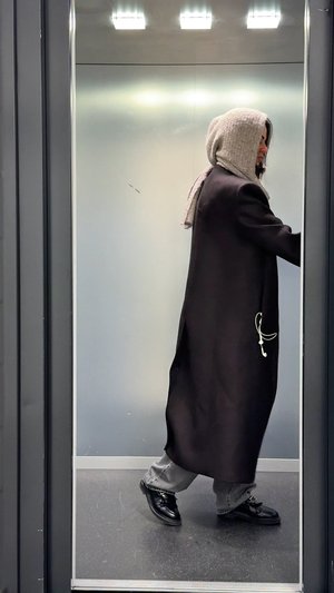 Persona con un lungo cappotto scuro e una sciarpa con cappuccio beige che cammina all'interno di un ascensore con pareti grigie e pavimento scuro.