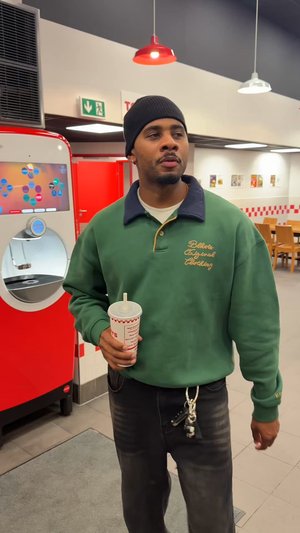 Sweat-shirt vert avec col bleu, texte brodé, pantalon noir, tenant une boisson avec une paille. En arrière-plan, un distributeur de boissons rouge.