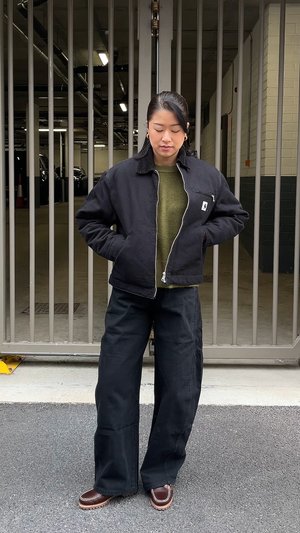 Veste noire à fermeture éclair portée sur un pull kaki, associée à un pantalon noir ample et des mocassins marron avec des coutures blanches.