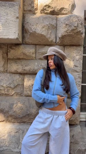Un sweat à zip bleu clair dans un matériel doux, associé à un pantalon de survêtement gris à jambes larges. Un chapeau beige et un sac à motifs viennent compléter l'ensemble. Fond en pierre.