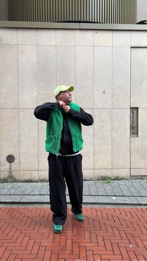 Zöld bolyhos mellény fekete kapucnis pulóver felett, fekete nadrággal és zöld cipővel párosítva. A háttérben egy textúrált kőfal és járda látható.