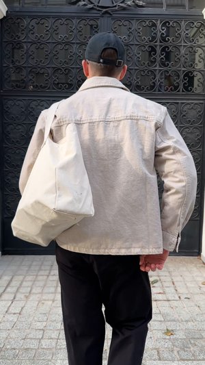 Homme portant une veste beige et une casquette noire, portant un grand sac fourre-tout off-white, debout sur un pavé en pierre devant une porte en fer décorative.
