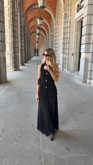 Top sans manches noir avec des boutons dorés, associé à une longue jupe noire. Accessoirisé avec un sac comportant des éléments ronds.