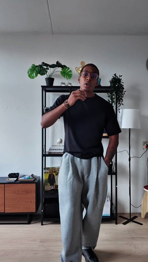 T-shirt noir à manches courtes et pantalon de survêtement gris clair portés par une personne, debout dans une pièce avec des plantes et une lampe en arrière-plan.