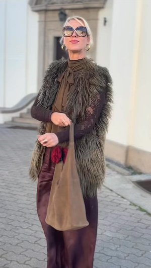 Faux fur gilet worn over a brown blouse, teamed with a long burgundy skirt. The person carries a beige bag embellished with a red tassel.
