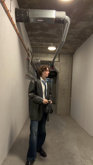 Veste noire en cuir, cravate bleue, chemise blanche et gants noirs portés par une personne dans un couloir en ciment avec tuyauterie et éclairage industriel.