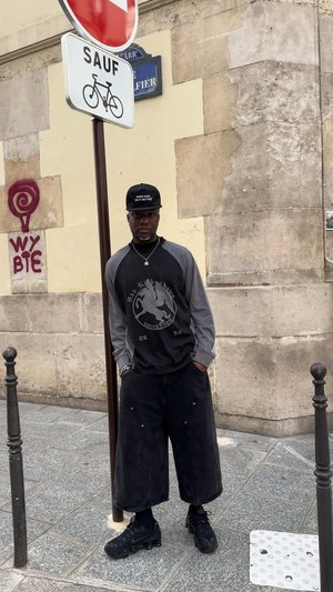 Schwarzes und graues Langarmshirt mit grafischem Design, schwarze weite Shorts, schwarze Sneakers und eine schwarze Kappe mit weißem Text. Städtisches Umfeld.